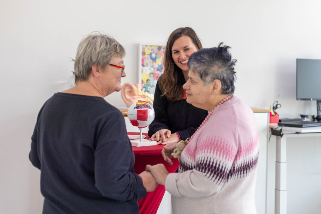 Drei Menschen sind in einer Gesprächssituation. Auf dem Stehtisch in der mitte des Bildes sieht man das Modell Auges und eines Ohrs. Die beiden Personen im Vordergrund halten sich freundschaftlich an der Hand und schauen sich an. Die Person im Hintergrund lächelt und schaut die Person im rechten Bildrand an.