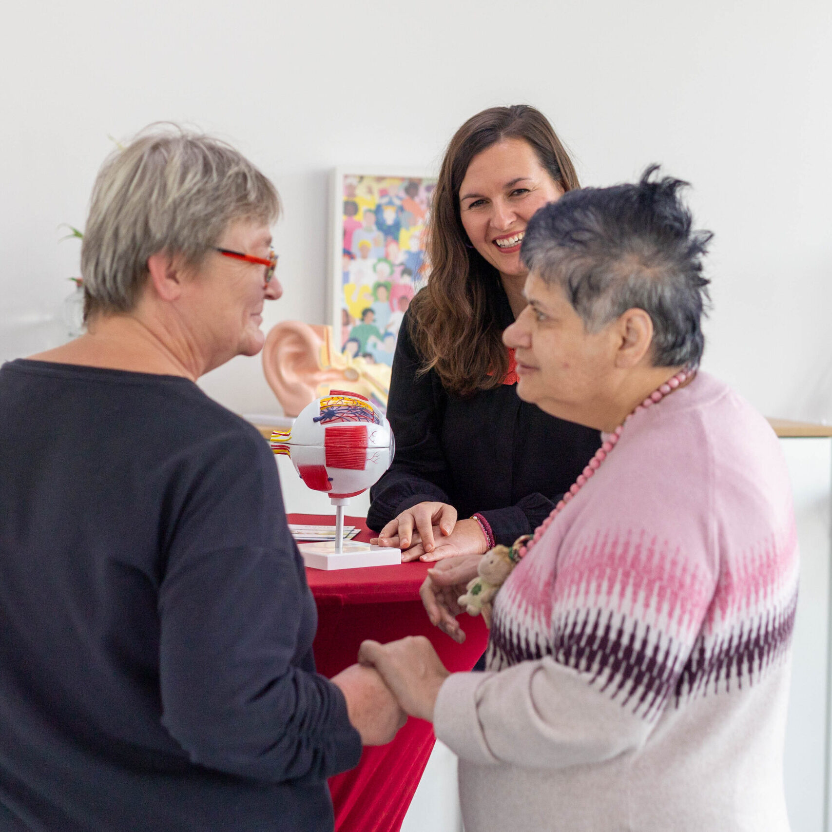 Drei Menschen sind in einer Gesprächssituation. Auf dem Stehtisch in der mitte des Bildes sieht man das Modell Auges und eines Ohrs. Die beiden Personen im Vordergrund halten sich freundschaftlich an der Hand und schauen sich an. Die Person im Hintergrund lächelt und schaut die Person im rechten Bildrand an.